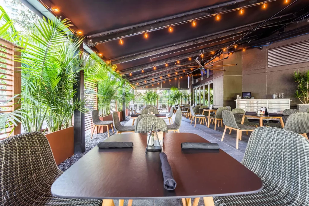 Covered patio with woven chairs, palm planters and string lights at Tap 42 Midtown Miami